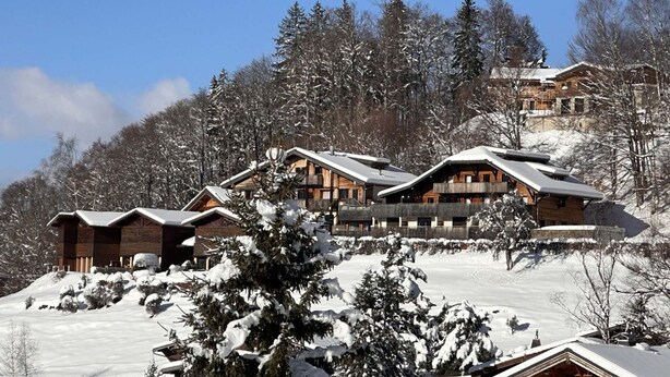 Eine Gruppe Holzhütten mit geneigten Dächern, umgeben von schneebedeckten Bäumen und einem klaren, blauen Himmel.