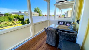 Ein Balkon mit Korbmöbeln, einem kleinen Tisch und Blick auf einen Garten und Häuser.