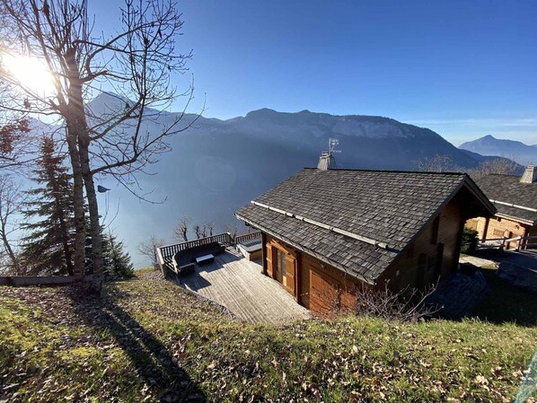 A wooden cabin with a sloped roof, a deck, and a view of mountains and a lake.
