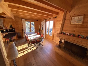A cozy cabin interior with a wooden dining table, chairs, a fireplace, and a view of the outdoors through large windows.