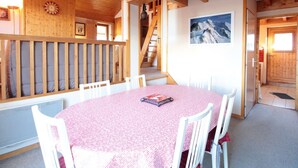 Ein gemütliches Zimmer mit rundem Tisch, weißen Stühlen, Holzbalken, einer Treppe und einem Poster mit Bergblick.