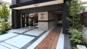 The entrance of a hotel with a tiled facade, a covered entrance, and a small garden area with plants. - Hotel Trend Omiya (Saitama)