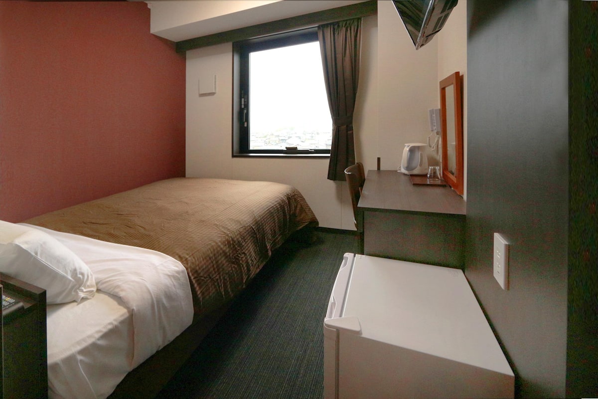 A hotel room with a bed, a desk, a chair, a window with curtains, and a view of the outdoors.