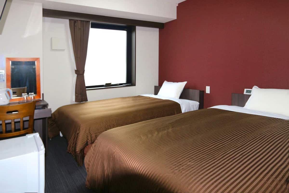 A hotel room with two beds, a desk with a computer, a window with curtains, and a red wall.