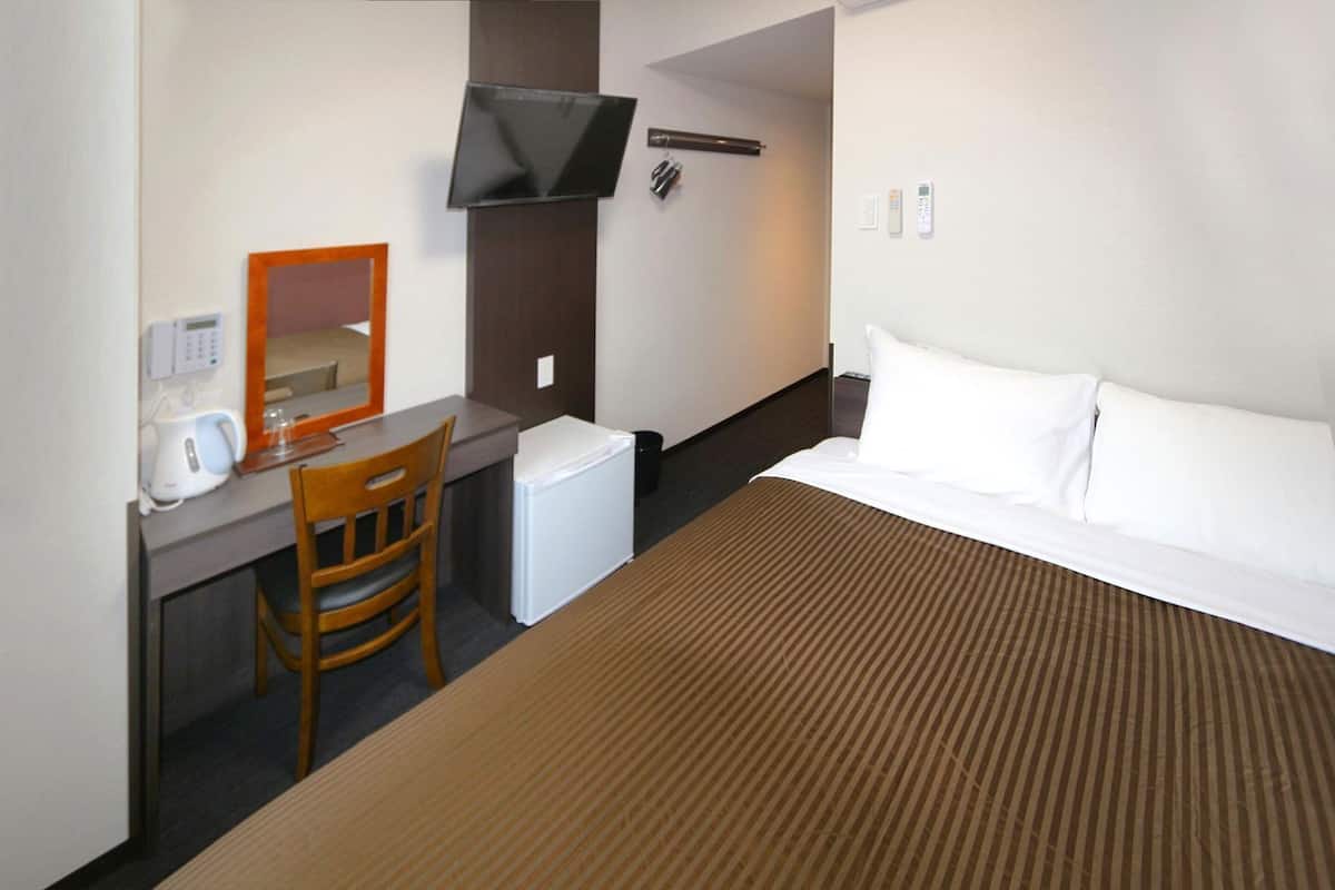 A hotel room with a bed, a desk with a chair, a television mounted on the wall, and a small refrigerator.