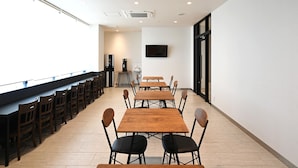A spacious dining area with multiple wooden tables and chairs, a large window providing natural light, and a television mounted on the wall. - ARIGATO STAY SUZUKA (Suzuka)