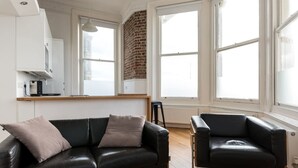 A modern living room with a black leather sofa and armchair, a kitchen area with white cabinets, and large windows with a brick wall. - Immaculate 1-bed Apartment on Hove Seafront (Hove)