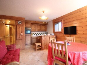 A cozy cabin-style room with wooden walls, a kitchen area with a stove, a dining table with chairs, and a television mounted on the wall. - 3-room cabin apartment with parking, near slopes and shops (Les Carroz d'Arâches)