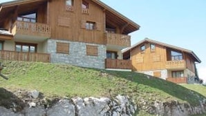 A wooden chalet with multiple balconies, stone walls, and a grassy hillside. - 3-room cabin apartment with parking, near slopes and shops (Les Carroz d'Arâches)