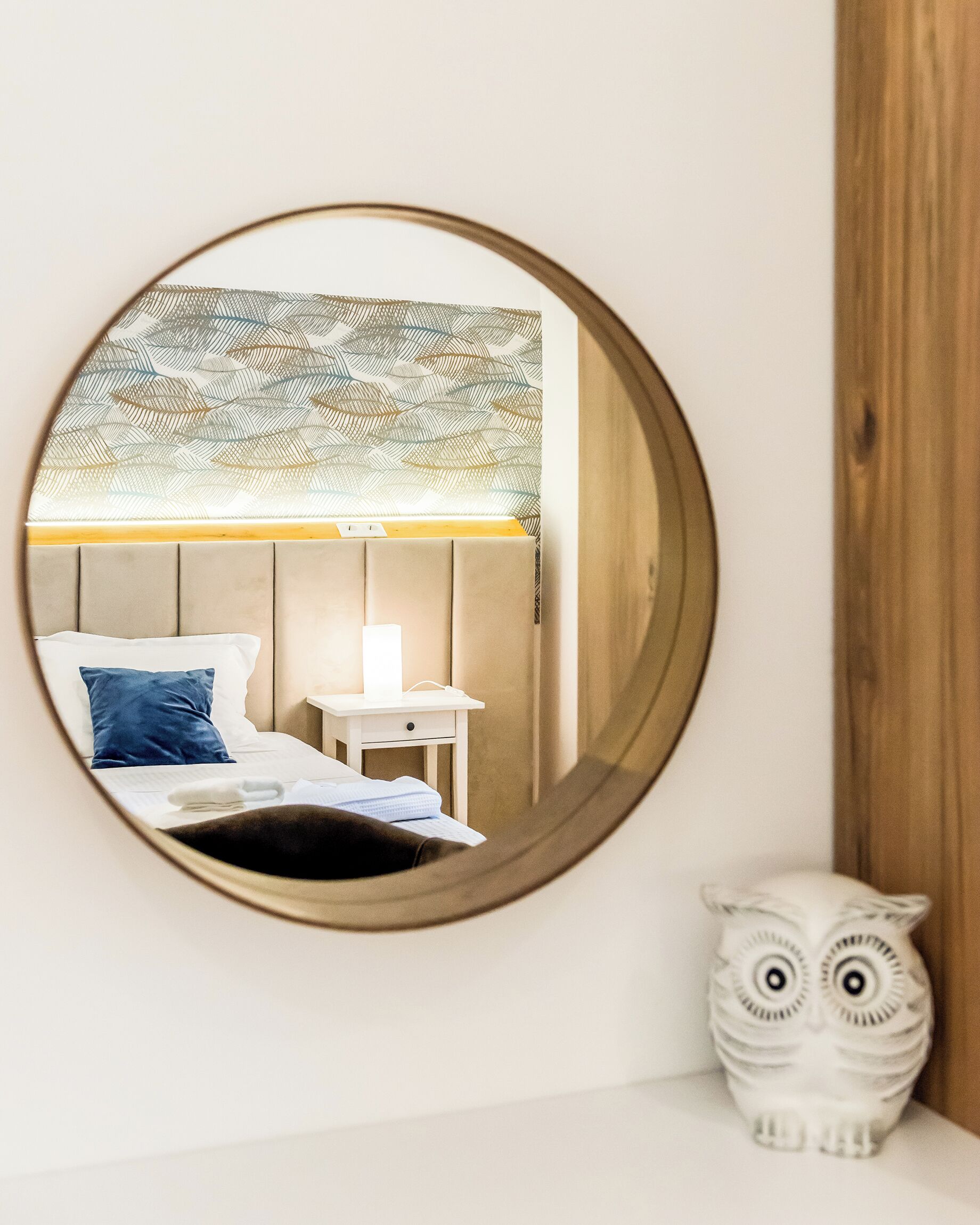 A round mirror reflecting a bed with a headboard, a bedside table with a lamp, and a white owl figurine.