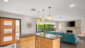 A modern kitchen with wooden cabinets and a central island, a living area with teal sofas and a stone fireplace, and a large TV mounted on the wall. - Mountain Views, Pool and Near Downtown (Palm Springs)