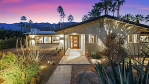 A modern house with a well-lit entrance, surrounded by desert plants and palm trees, with mountains in the background. - Mountain Views, Pool and Near Downtown (Palm Springs)