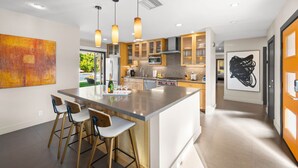A modern kitchen with a central island, bar stools, pendant lighting, and artwork on the walls. - Mountain Views, Pool and Near Downtown (Palm Springs)