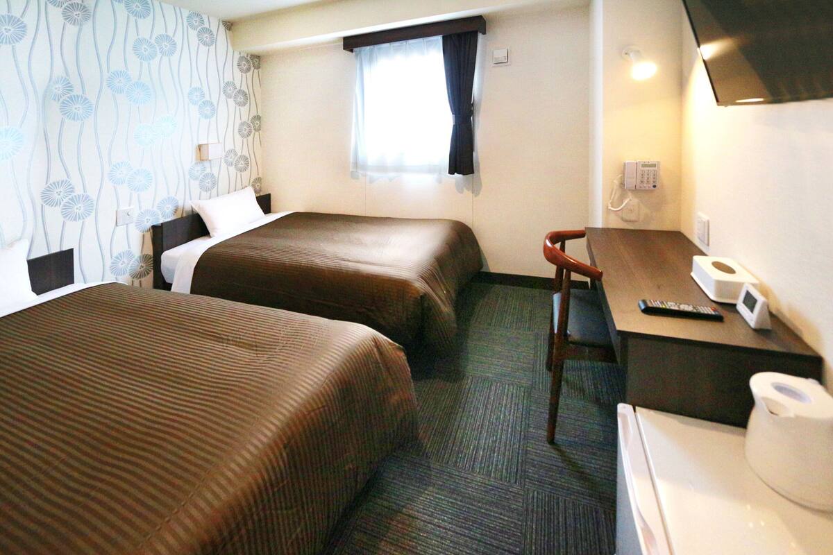 Ein Hotelzimmer mit zwei Betten, einem Schreibtisch, einem Sessel, einem Telefon und einem fenster mit Vorhängen.