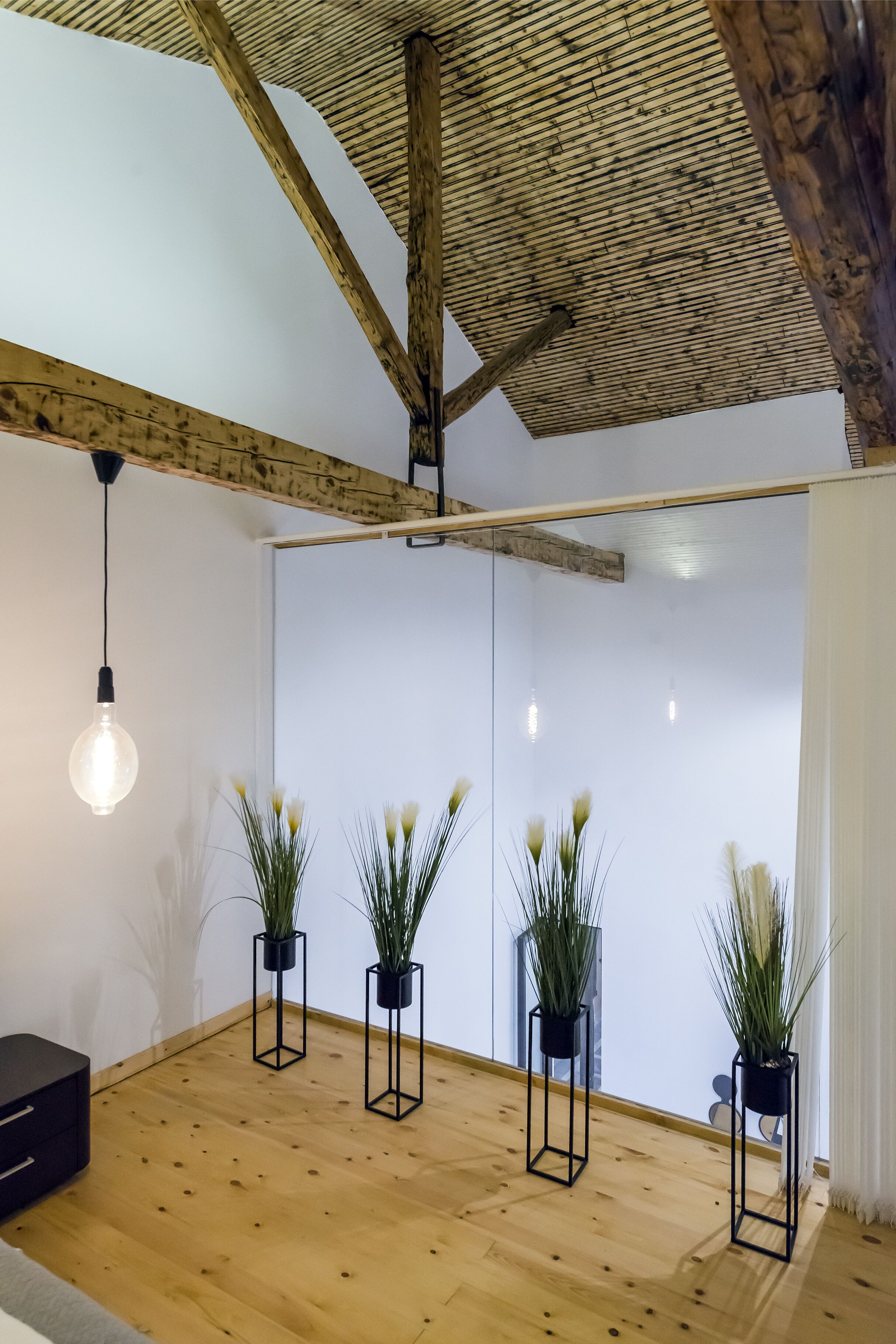 A room with wooden flooring, a sloped ceiling with exposed beams, and four black square planters with plants.