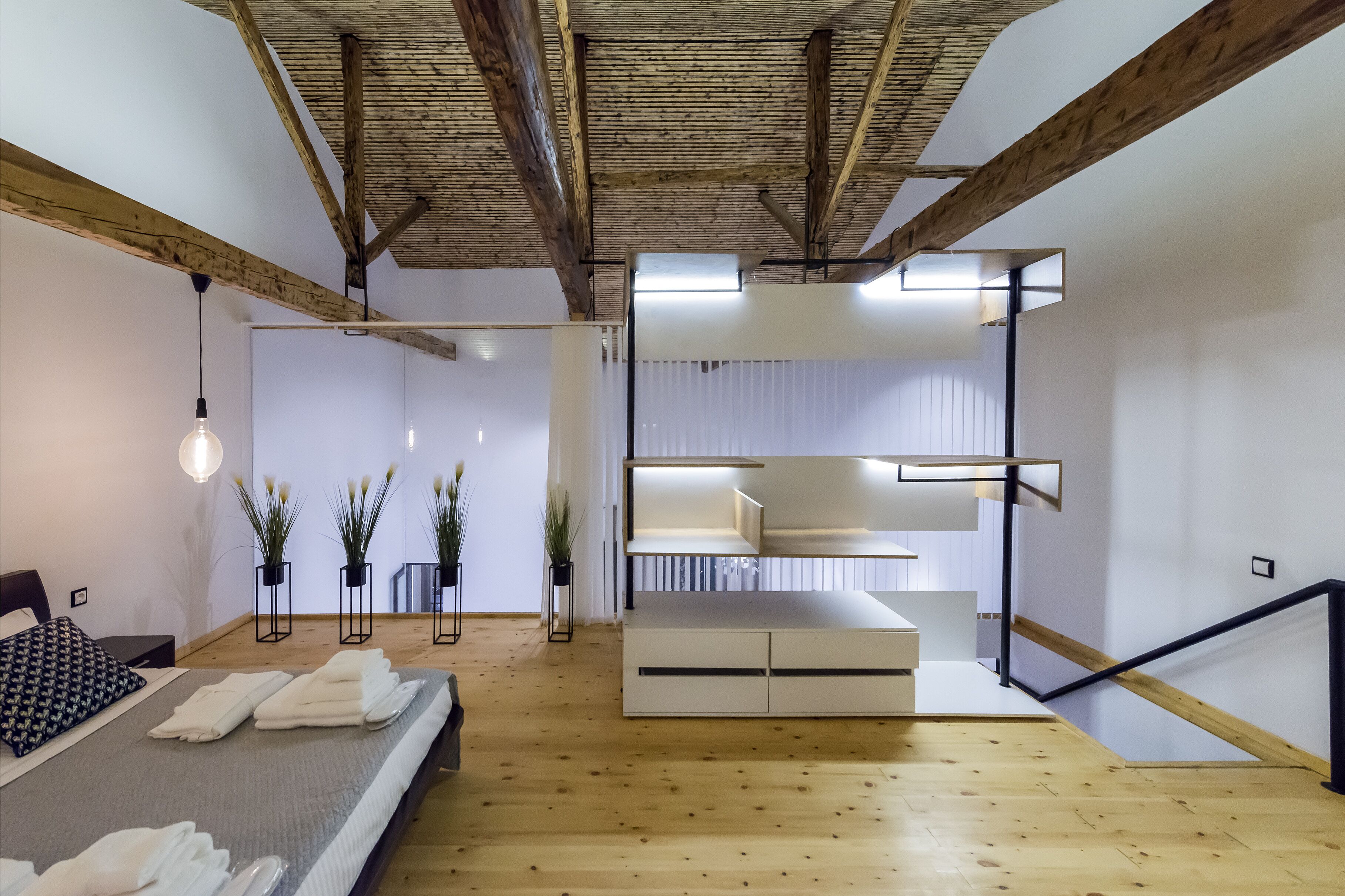 A modern hotel room with a wooden floor, a bed with neatly folded towels, a bedside table, and a built-in bench with storage drawers.