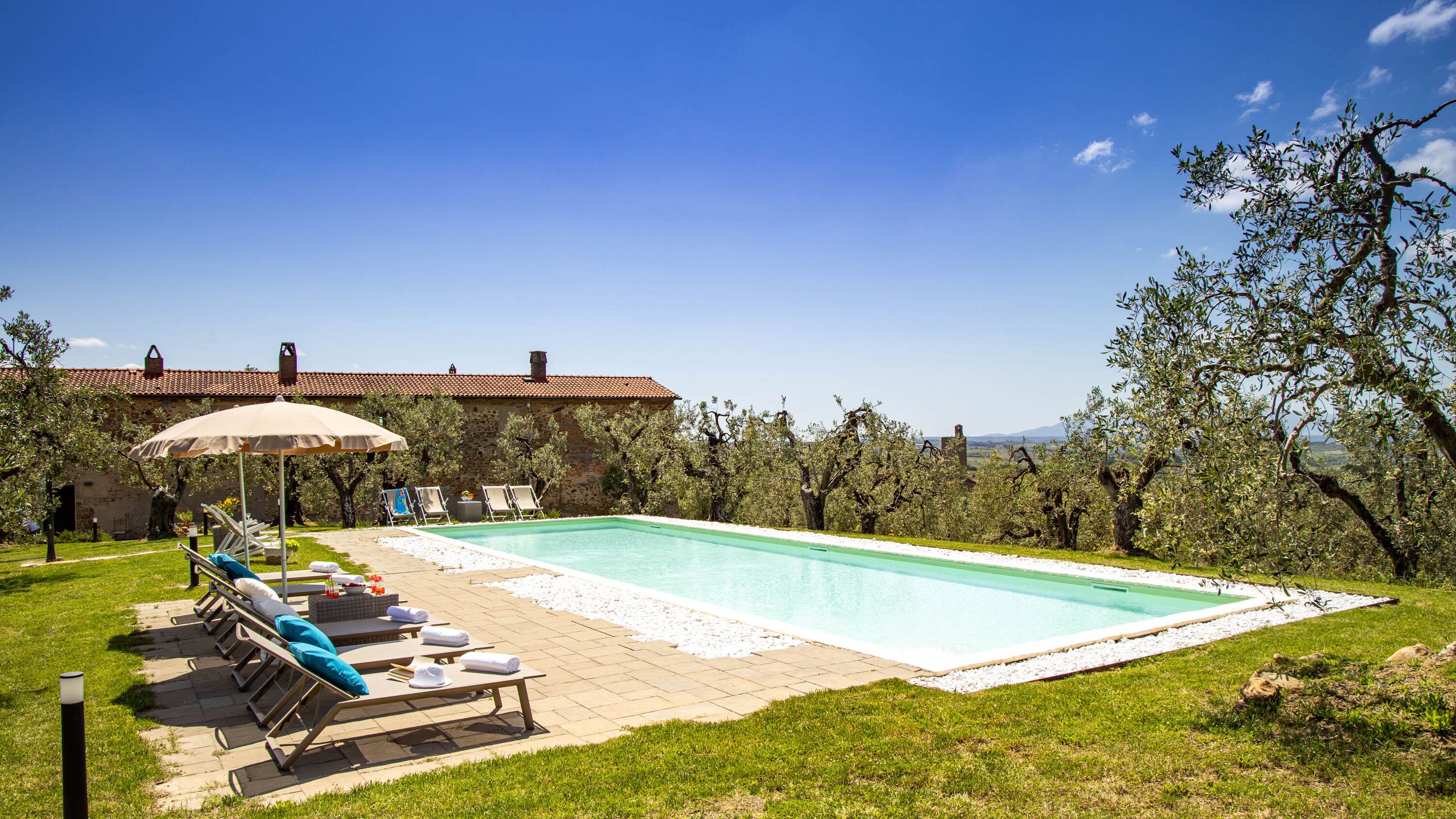 A swimming pool with lounge chairs and an umbrella, surrounded by trees and a stone building.