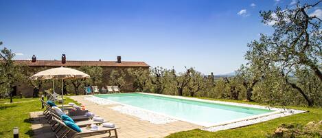 A swimming pool with lounge chairs and an umbrella, surrounded by trees and a stone building.
