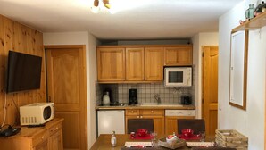 Eine kompakte Küche mit Holzschränken, einer Mikrowelle, einer Kaffeemaschine und einem Geschirrspüler. Ein Esstisch mit Stühlen und ein an der Wand befestigtes Fernsehgerät sind vorhanden.