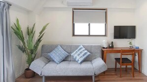 A living room with a grey sofa, two patterned cushions, a wooden desk, a television, a potted plant, and a window with blinds. - Dandelion (Tokyo)