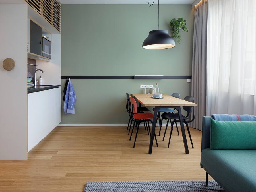 A modern living room with a sofa, a dining table with chairs, and a kitchen area with cabinets and a hanging plant.