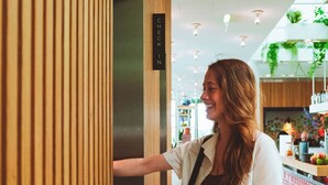 A person standing in a room with vertical wooden slats, a sign that reads 'CHECK IN', and a counter with plants and items in the background. - Zoku Vienna (Vienna)