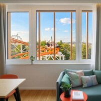 Quarto com uma grande janela com vista para uma montanha-russa, um sofá com almofadas, uma mesa pequena com um livro e uma planta em vaso.