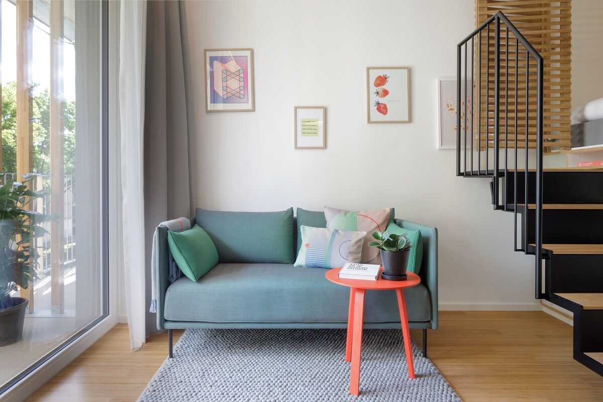 A modern living room with a teal sofa, a small orange table, and a staircase with black railings.