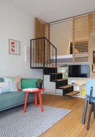 Um espaço de estar moderno com um sofá verde, uma pequena mesa com uma planta, uma televisão e uma escada com persianas de madeira.