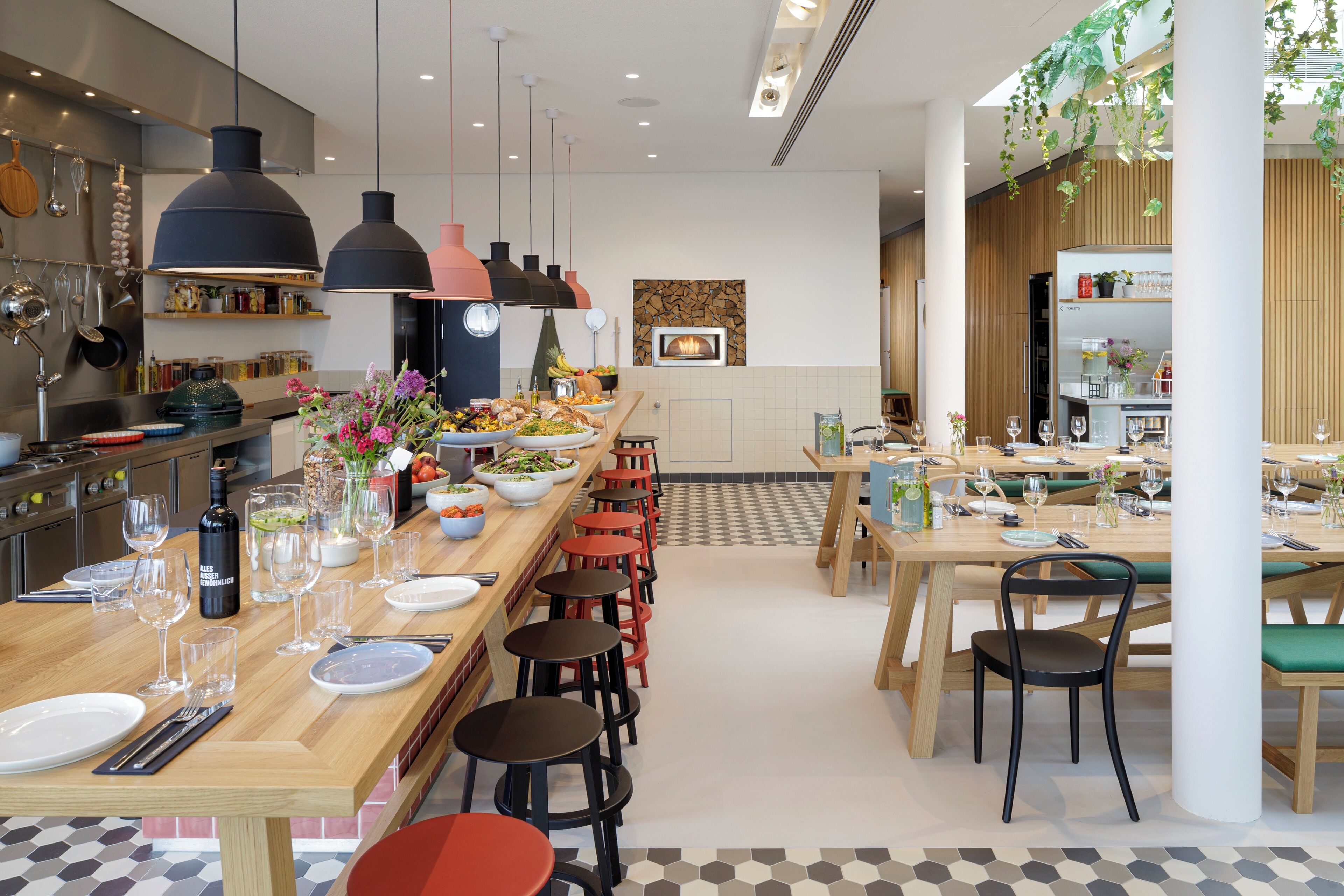 A modern restaurant with a long communal table, multiple seating options, and a central fireplace.
