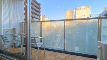Uma varanda com vista para a paisagem urbana, incluindo um edifício alto e uma grua. Há cadeiras de metal e uma mesa na varanda.