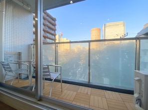 Uma varanda com vista para a paisagem urbana, incluindo um edifício alto e uma grua. Há cadeiras de metal e uma mesa na varanda.