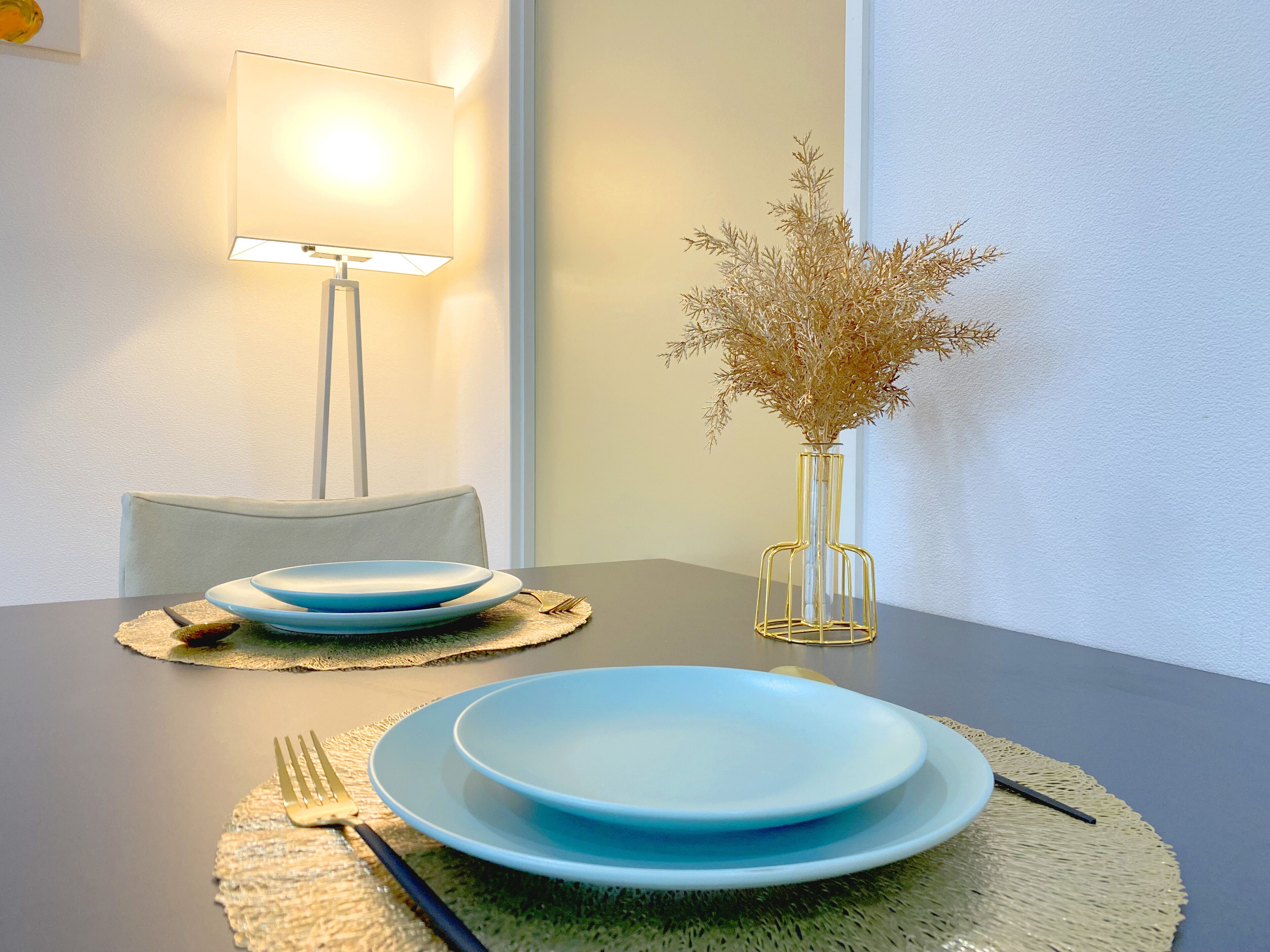 Een eettafel met blauwe borden, een gouden vork, een glazen vaas met gedroogde planten en een vloerlamp.