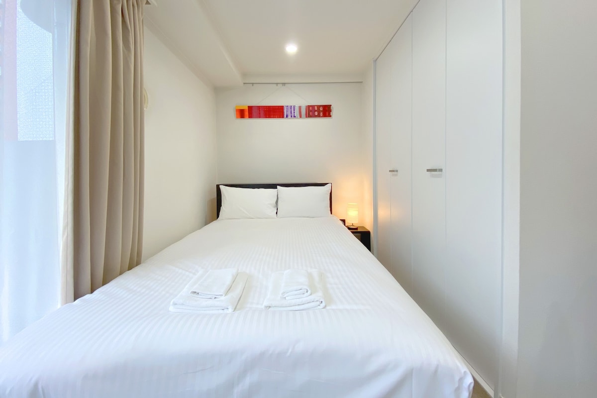 Cama bem arrumada com lençóis e toalhas brancas, luminária de cabeceira e janela com cortinas.