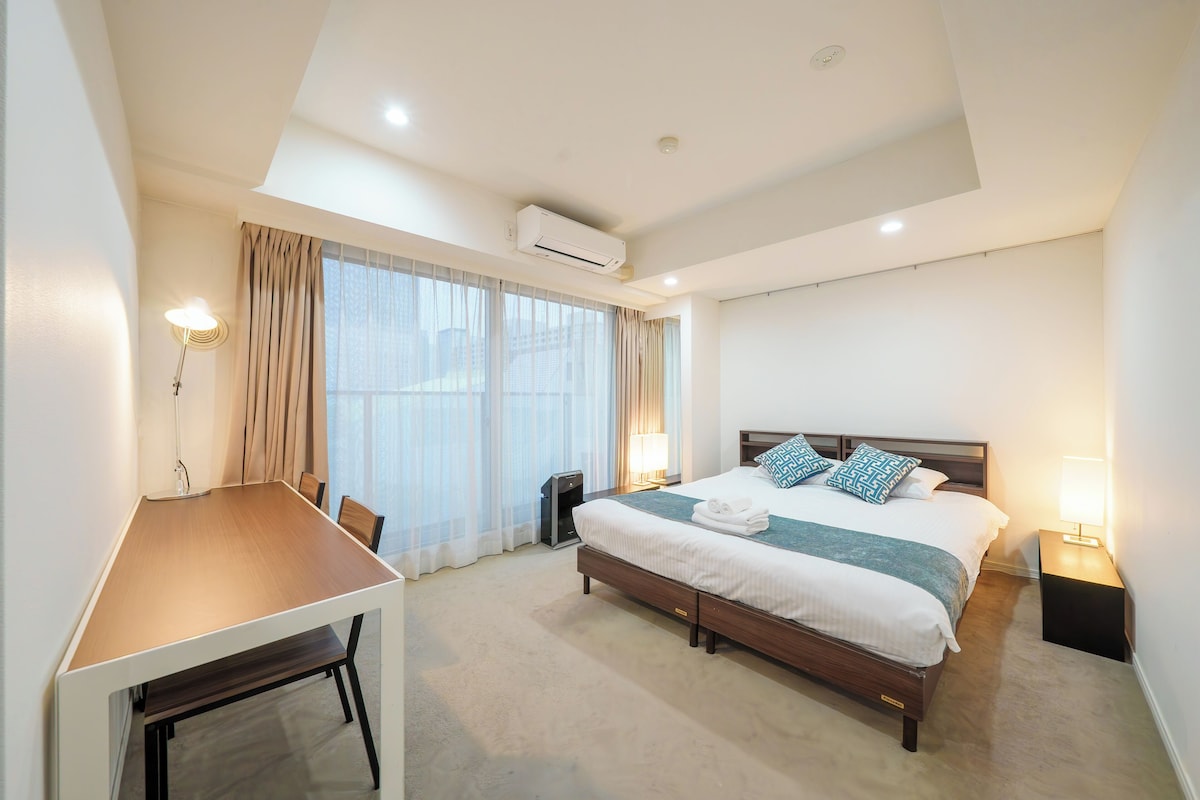 Quarto de hotel moderno com uma cama grande, mesas de cabeceira, uma escrivaninha e uma janela ampla com cortinas.