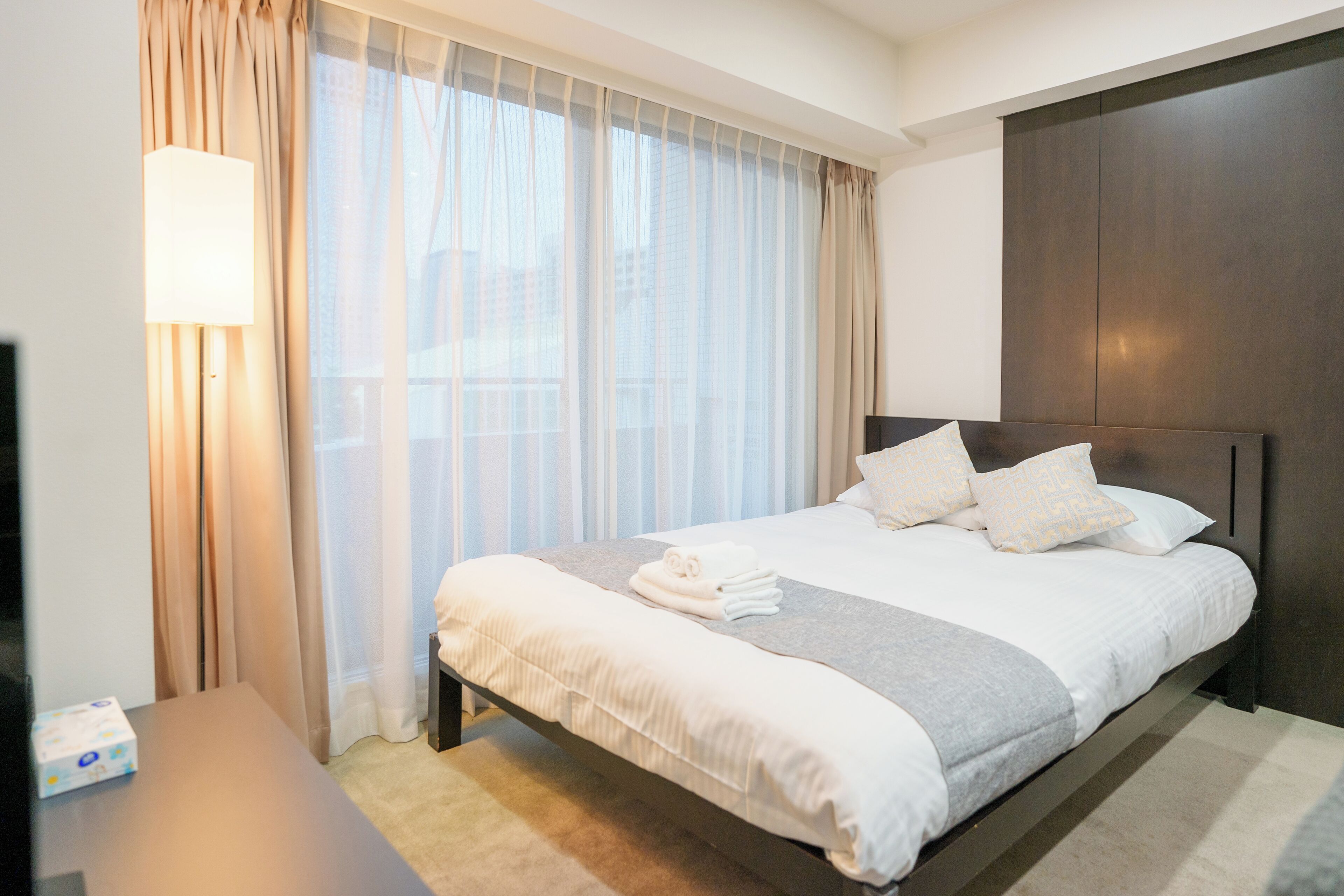 A hotel room with a bed, a bedside table, a lamp, and a view of the city through sheer curtains.