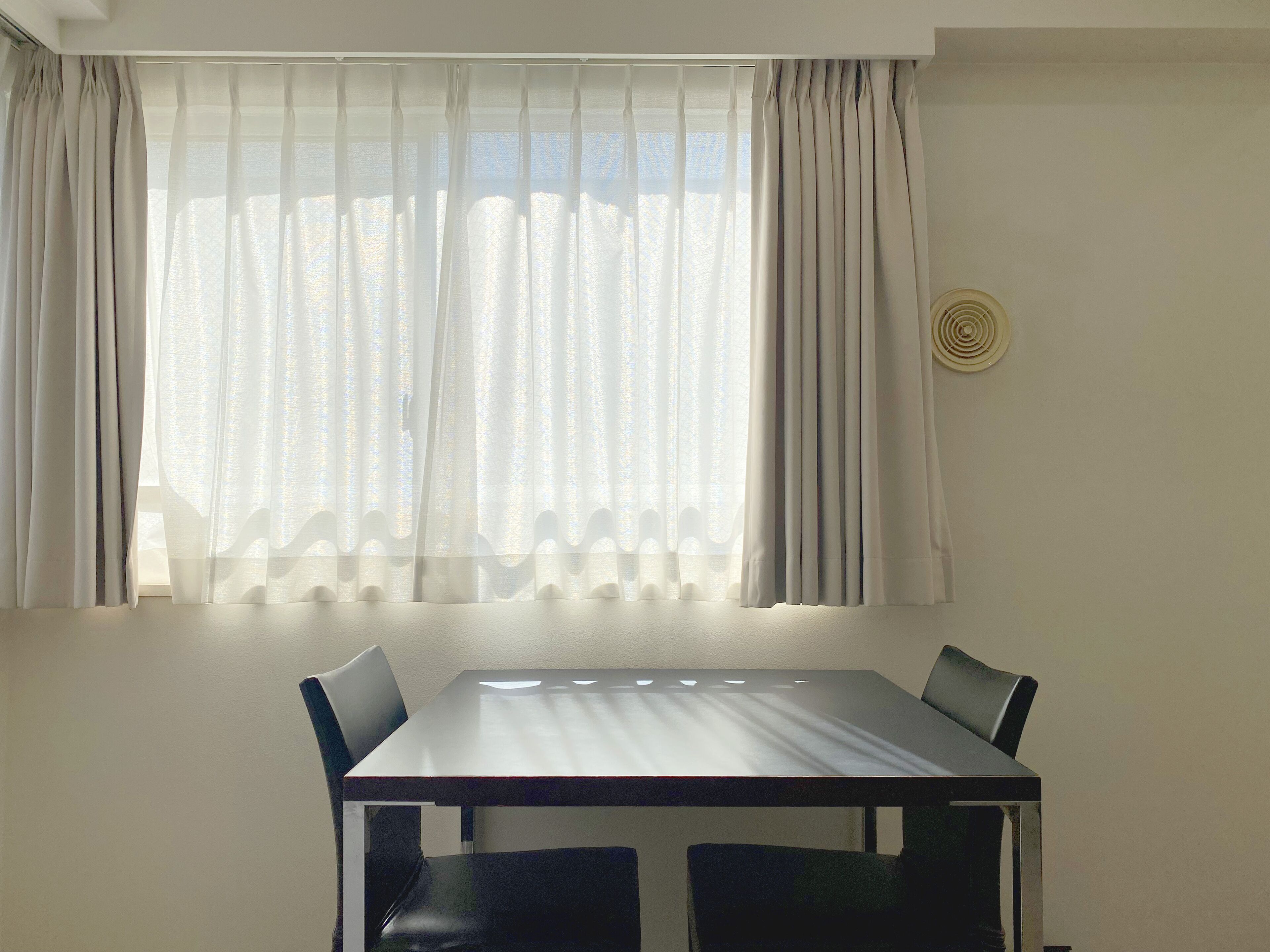 Uma área de refeições com mesa e cadeiras, uma janela com cortinas leves e um ar-condicionado fixado na parede.