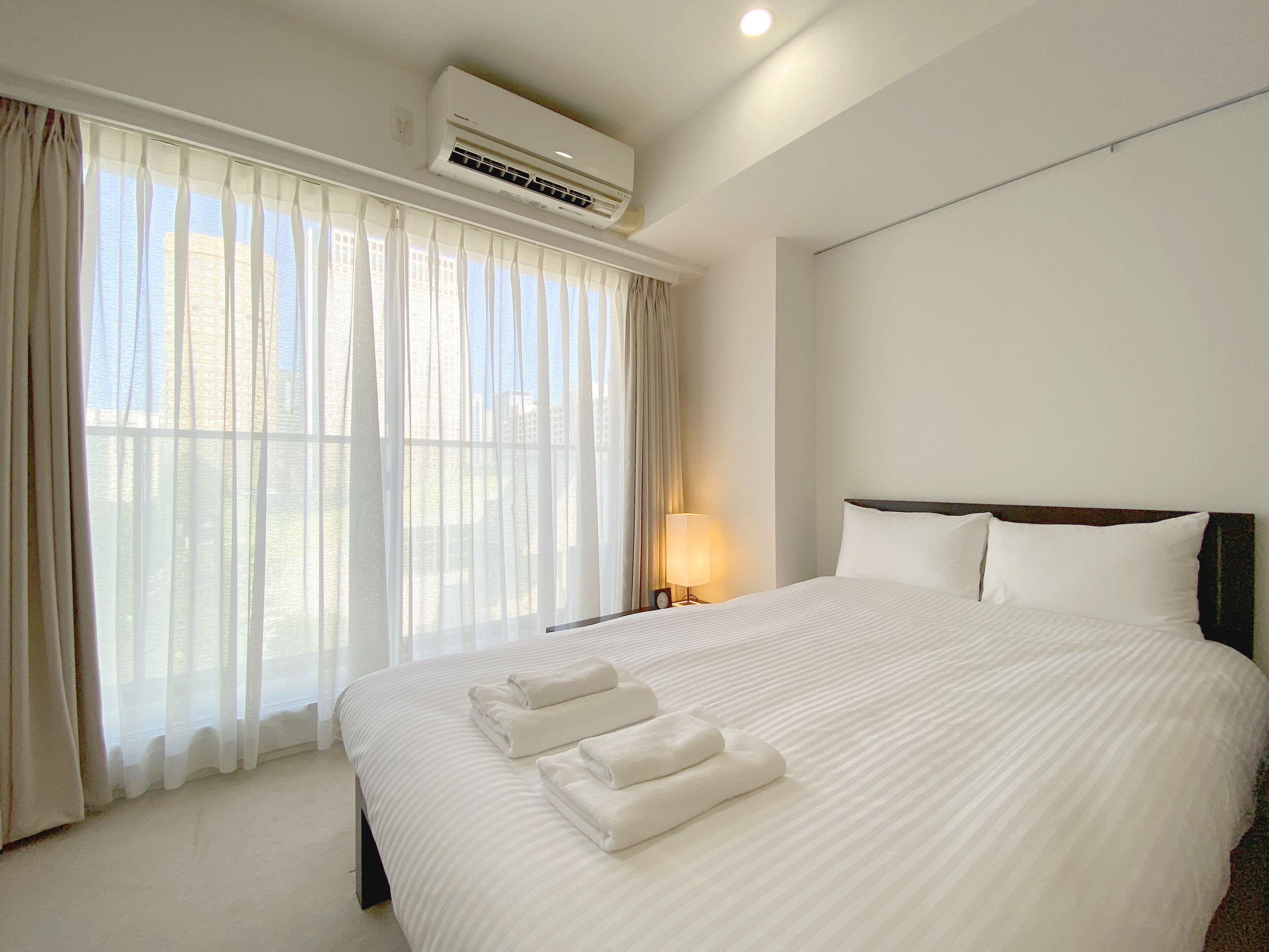 A hotel room with a large bed, white linens, a bedside lamp, and an air conditioning unit.