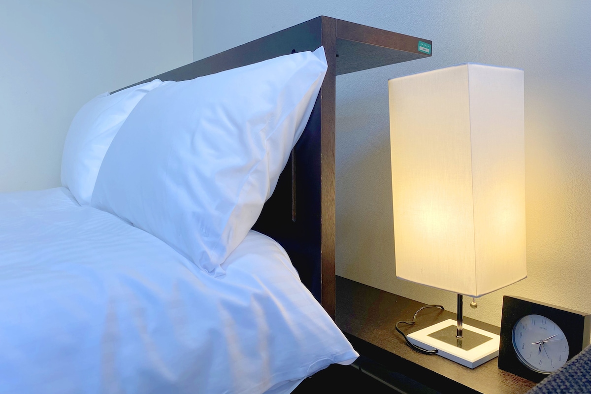 Uma cama bem arrumada com lençóis brancos, uma mesa de cabeceira com abajur e um relógio.