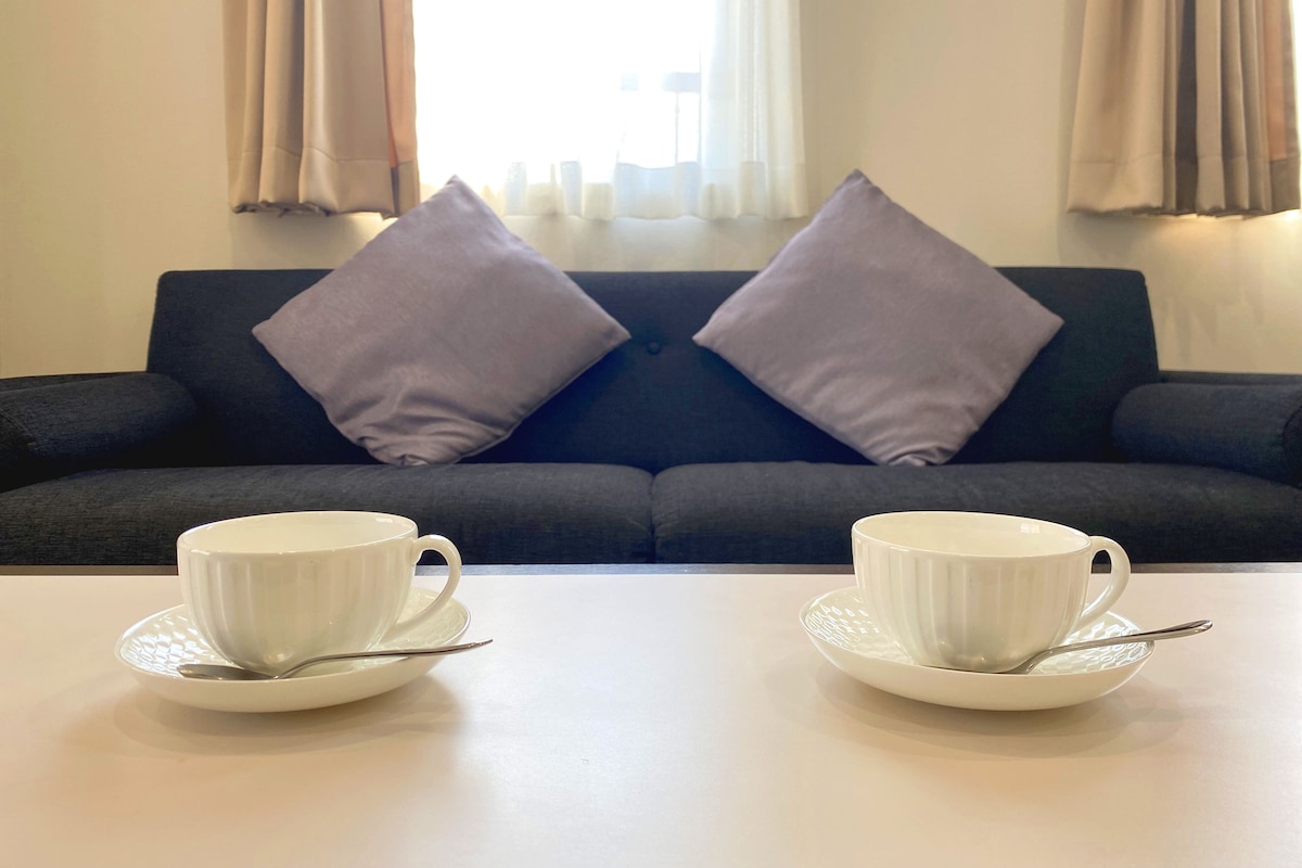 Quarto com um sofá escuro, duas chávenas brancas em pires e uma janela com cortinas.