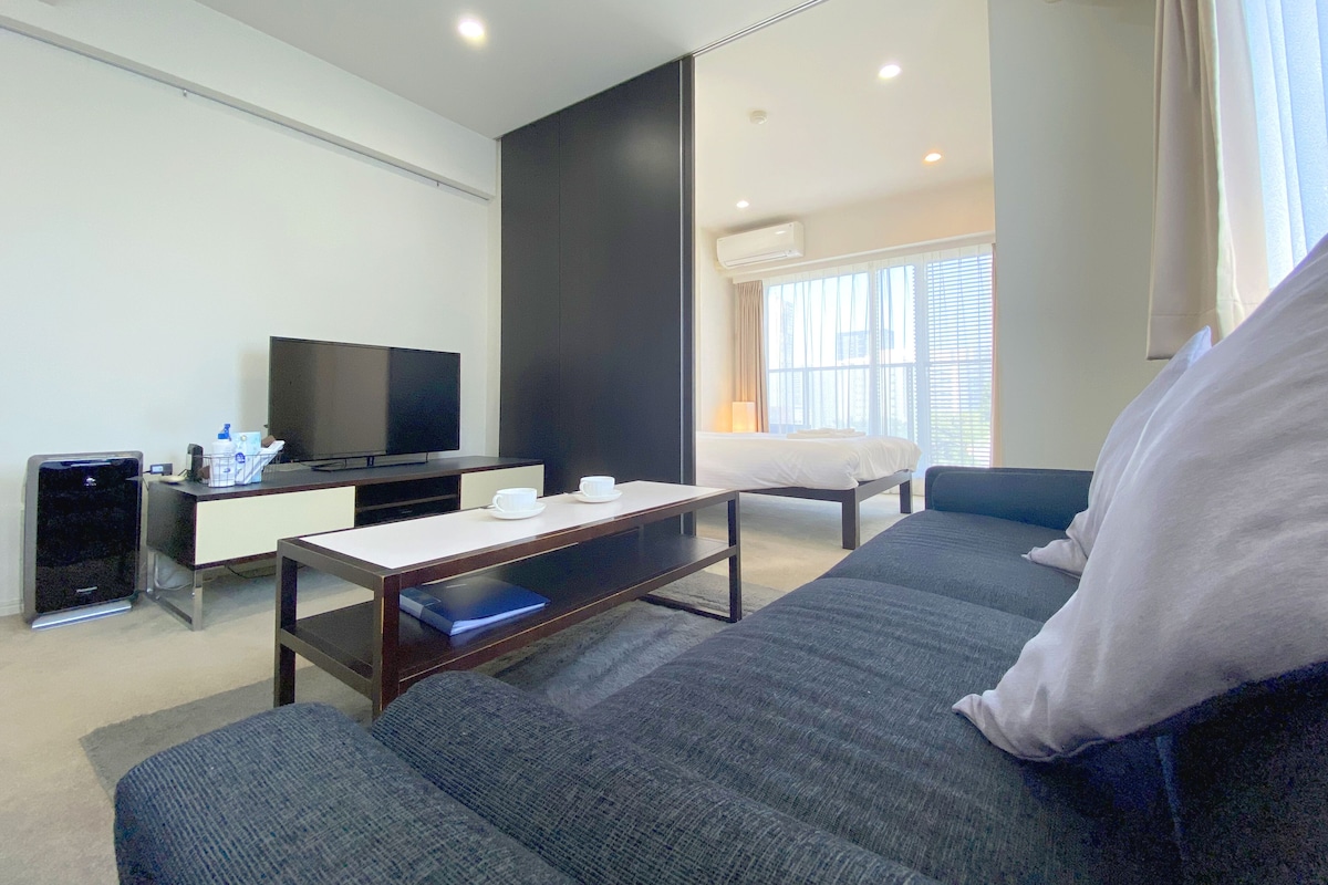 Quarto de hotel moderno com uma cama grande, um sofá, uma TV, uma mesa de trabalho e uma janela com cortinas.