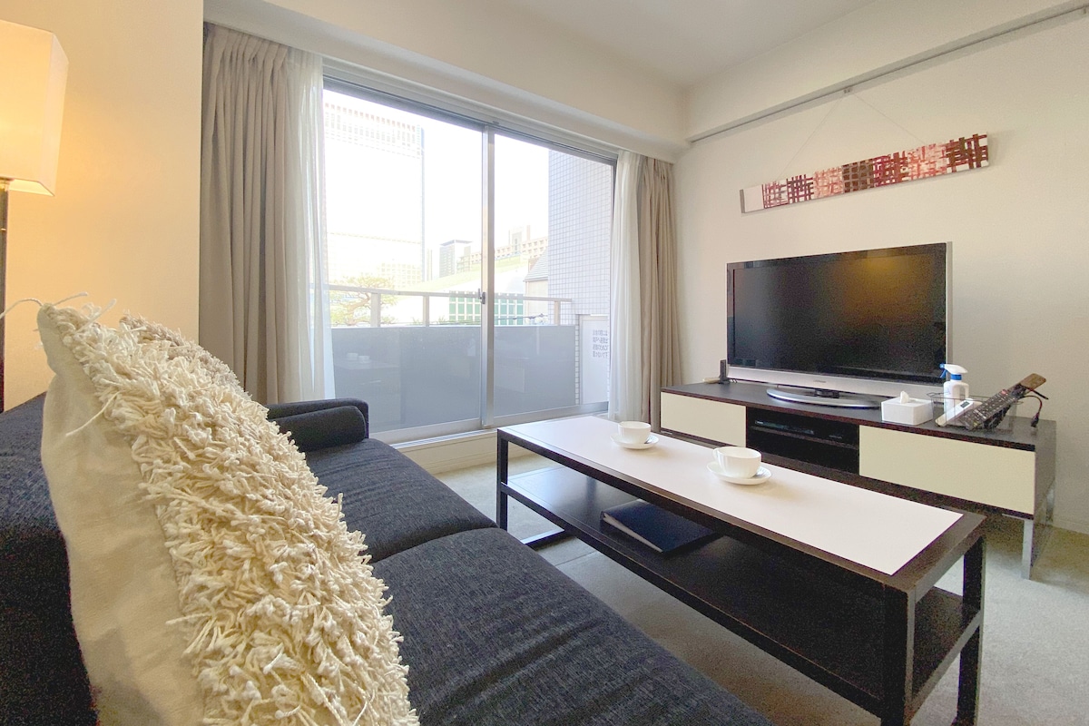 Quarto de hotel moderno com uma janela grande, uma televisão de tela plana, um sofá, uma mesa de centro e uma pequena escrivaninha com abajur.