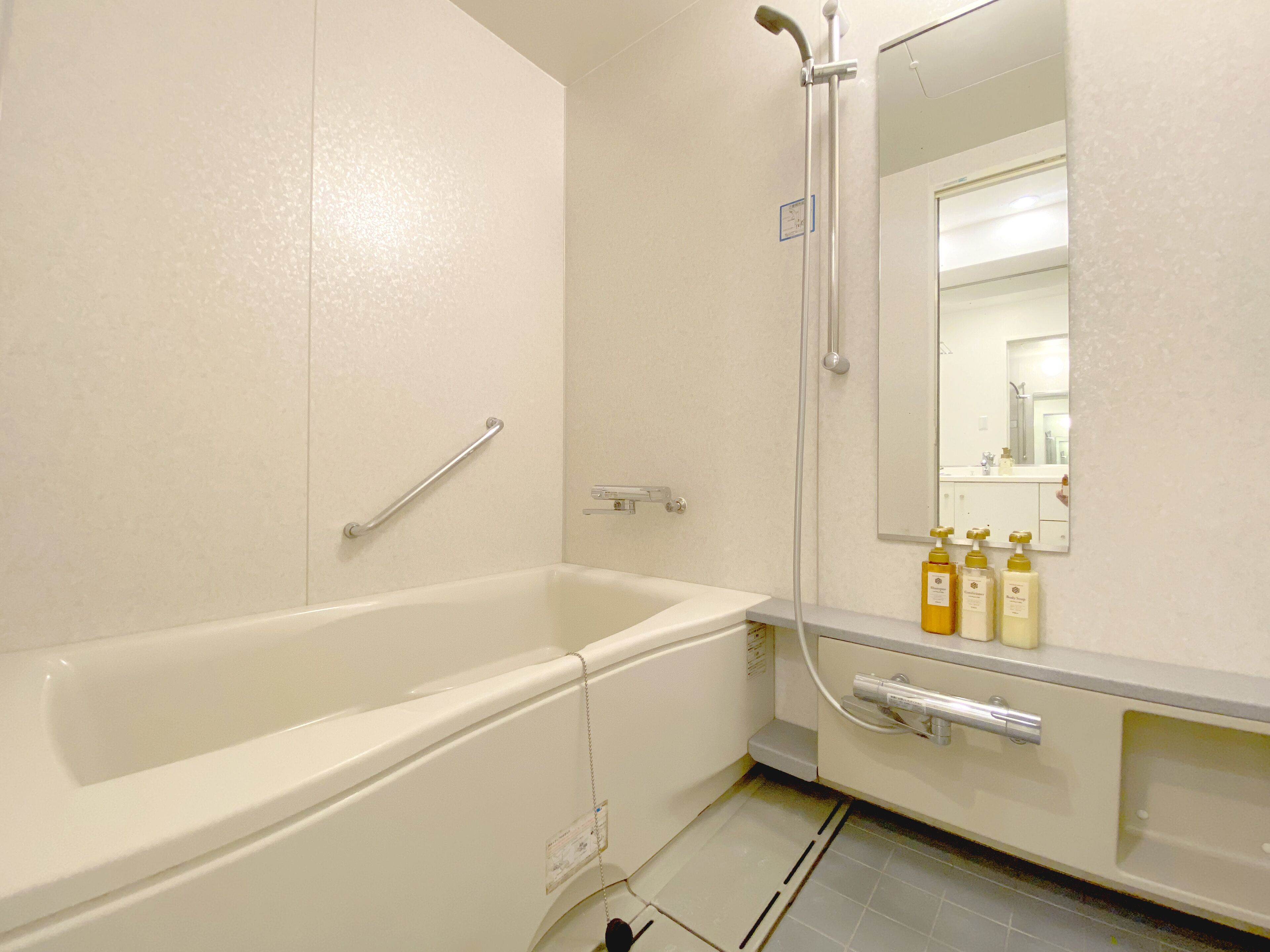 A bathroom with a bathtub, a shower, a handrail, a mirror, and toiletries on the counter.