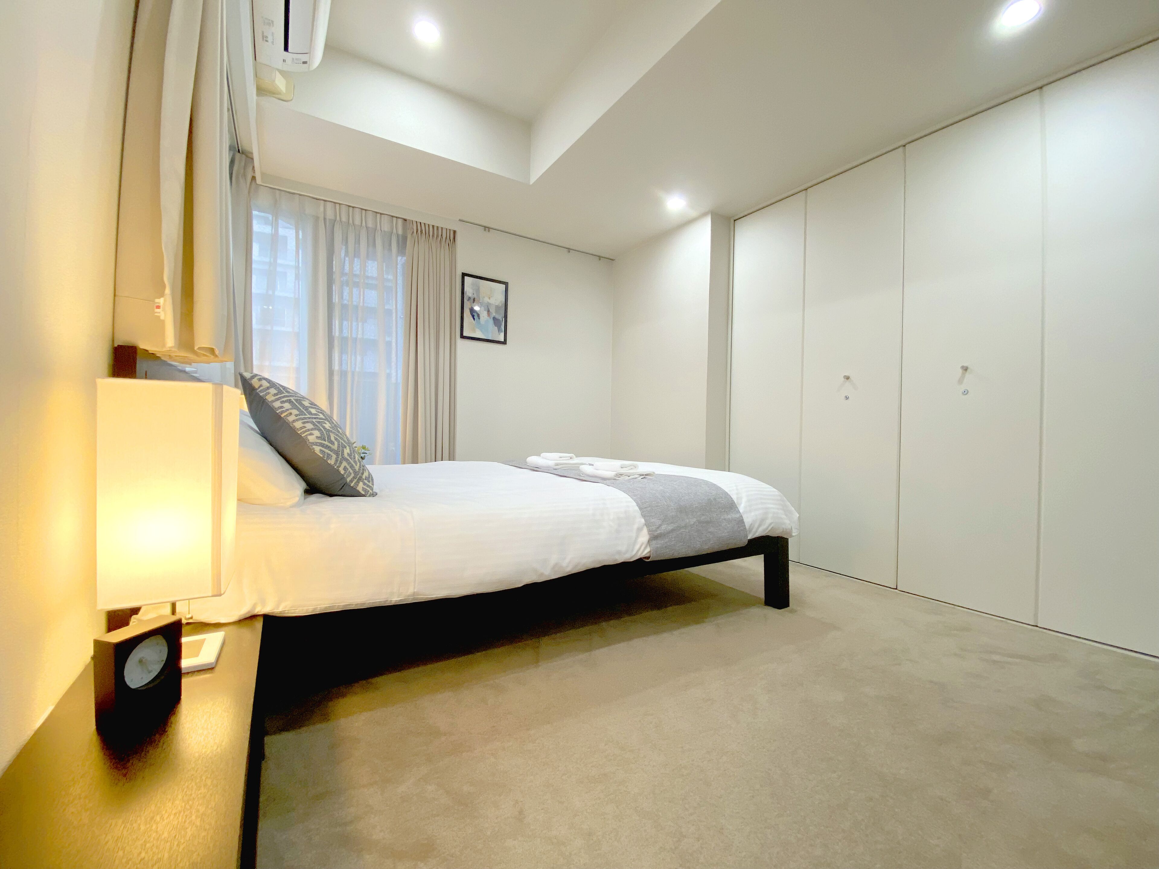 A bedroom with a bed, a bedside lamp, a clock, a window with curtains, and a wardrobe.
