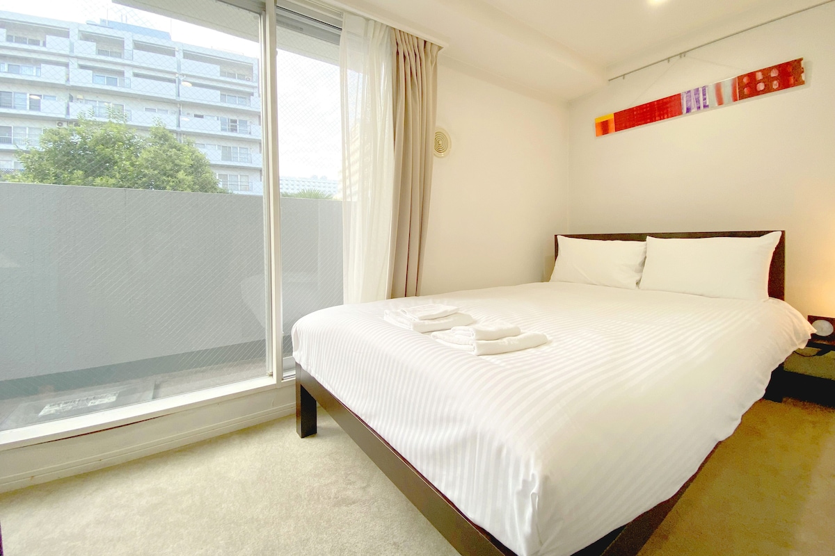 Quarto de hotel com uma cama grande, janela com cortinas e vista para um edifício, além de um quadro abstrato em vermelho e branco na parede.