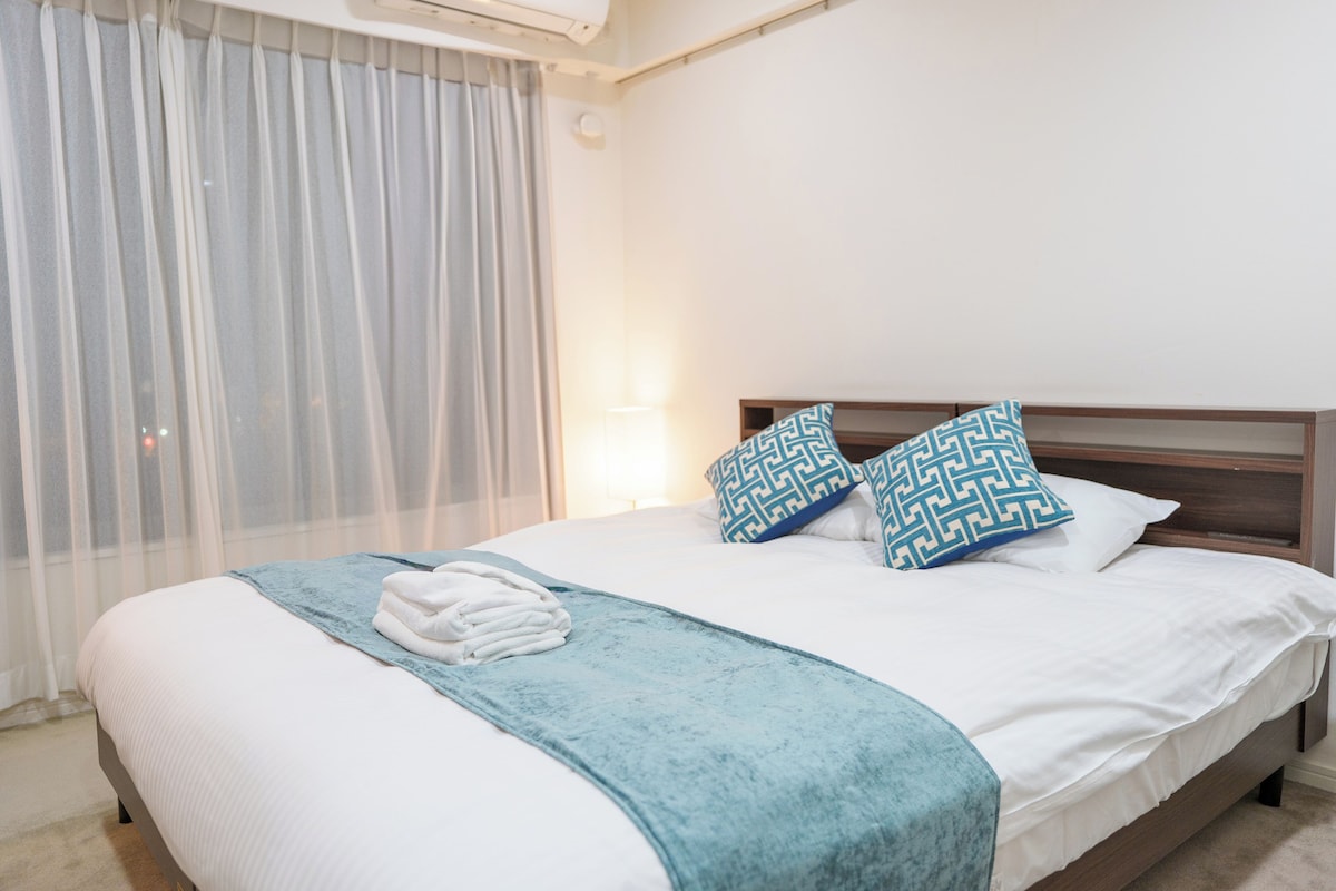 Cama bem arrumada com manta azul e duas almofadas estampadas, uma toalha branca dobrada na cama, e uma janela com cortinas leves.