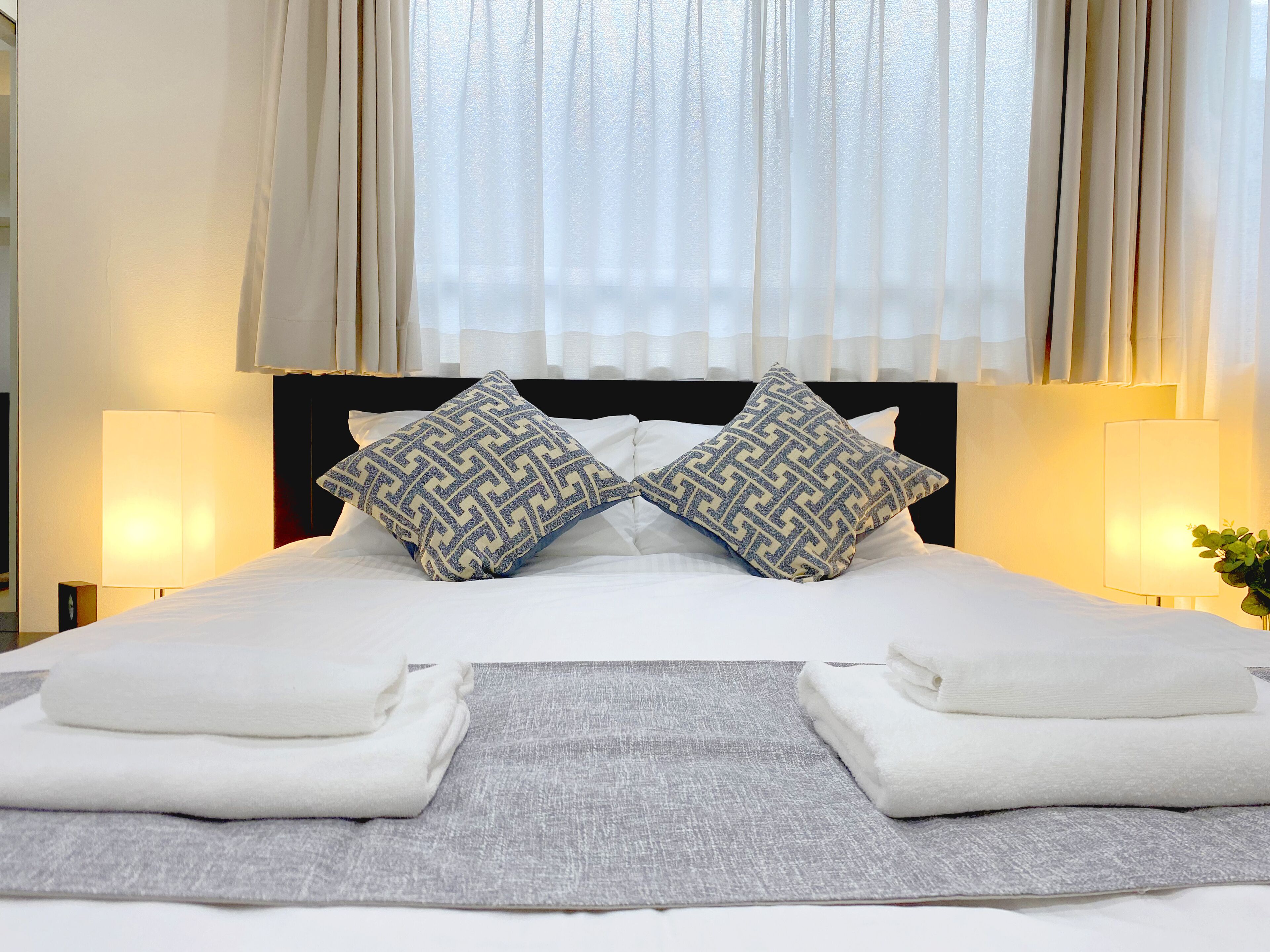 Un letto rifatto con due cuscini decorati, una coperta grigia e due asciugamani piegati.
