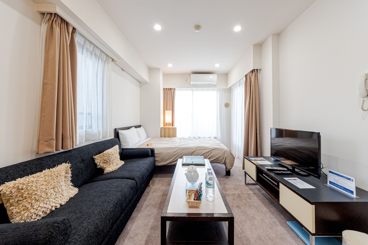 Quarto de hotel com sofá preto, uma cama, escrivaninha com televisão e janela com cortinas.