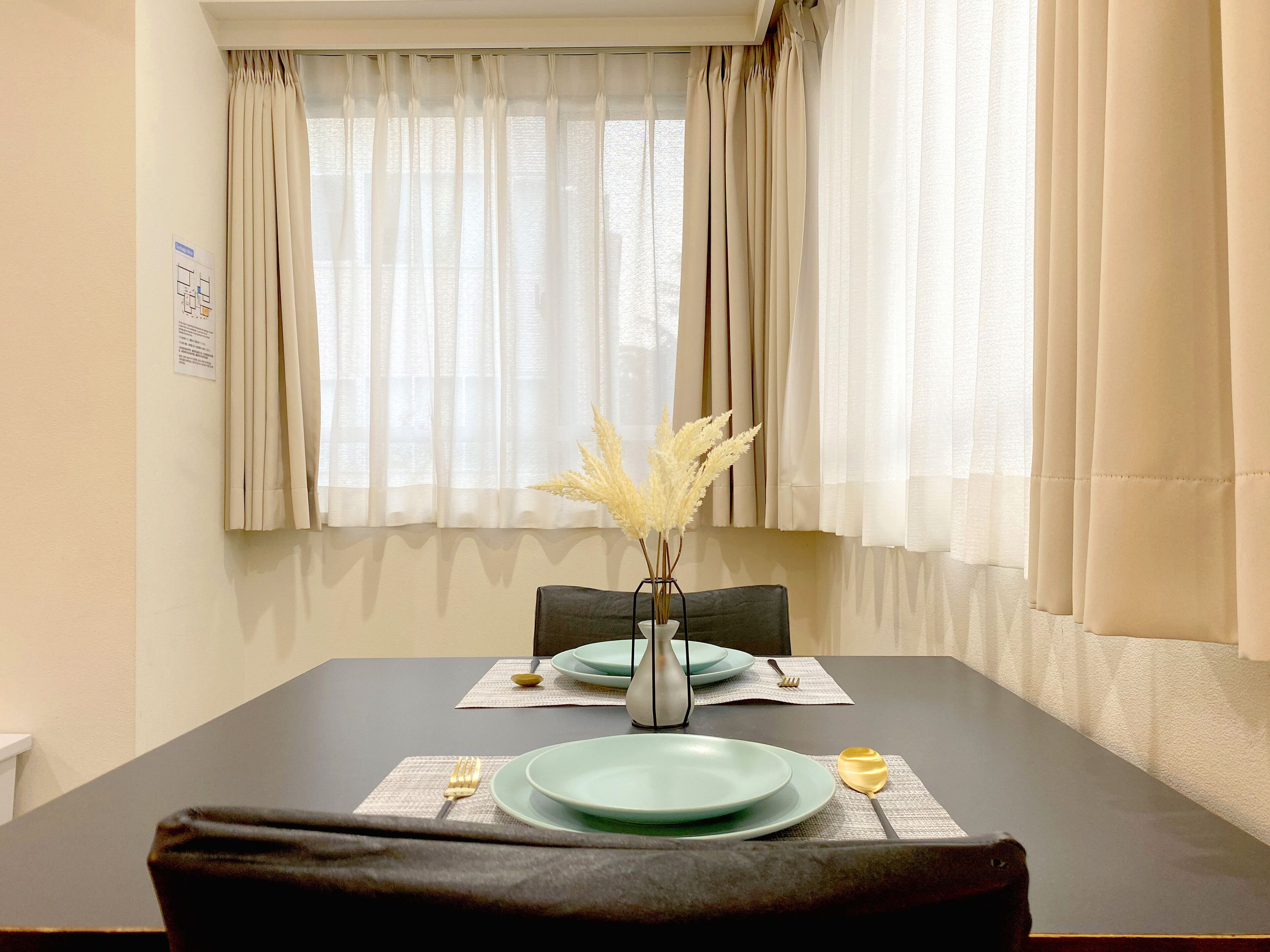 Un coin repas avec une table dressée pour deux personnes, un vase contenant des plumes de jarre séchées et une fenêtre ornée de rideaux légers.