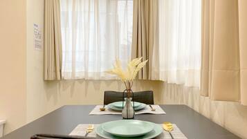 Un coin repas avec une table dressée pour deux personnes, un vase contenant des plumes de jarre séchées et une fenêtre ornée de rideaux légers.